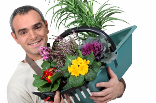 The image shows a man smiling and holding a teal garden tool caddy filled with a variety of colorful plants and flowers, including yellow, purple, and red blooms, in an outdoor garden setting. In the background, there is a lush green hedge and a well-maintained lawn with vibrant grass, edged with a flower bed containing soil and small shrubs. The garden appears tidy and landscaped, featuring a combination of lawn, borders, and small trees, reflecting typical features of a private outdoor space in Mayfair. The scene suggests outdoor gardening and maintenance activities, aligning with professional garden care services offered in the area. The weather appears bright and clear, with natural daylight illuminating the garden's greenery and floral accents, emphasizing a well-kept, attractive garden environment suitable for regular landscaping and lawn care services from Garden Maintenance Mayfair.