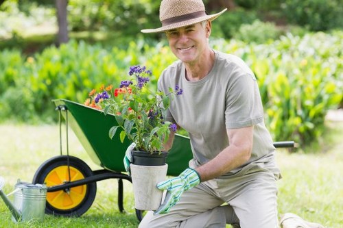 A gardener wearing a beige sunhat and light grey T-shirt is kneeling on a well-maintained lawn in a lush, green outdoor space, holding a potted flowering plant with purple, red, and pink blossoms. Behind him, there is a wheelbarrow with a bright yellow wheel, partially filled with gardening tools, and a watering can resting on the grass. The garden features a neatly trimmed hedge or row of shrubs in the background, along with tall trees providing shade in a natural, peaceful setting. The scene suggests a daytime environment with natural sunlight highlighting the vibrant colors of the plants and the greenery surrounding the area, indicating outdoor maintenance work typical of professional gardening services in the Mayfair area.